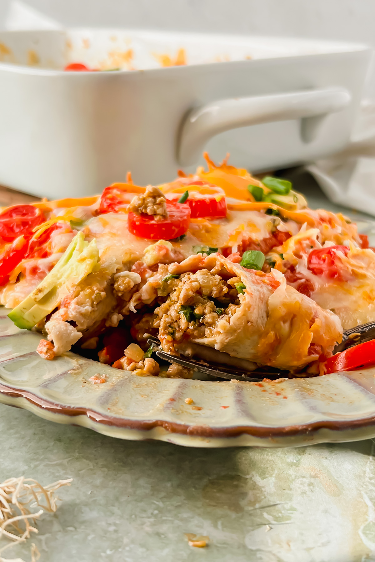 baked healthy ground turkey enchiladas garnished with cherry tomato slices, avocado slices, green onion on gray serving plate with pan of additional enchiladas in background.