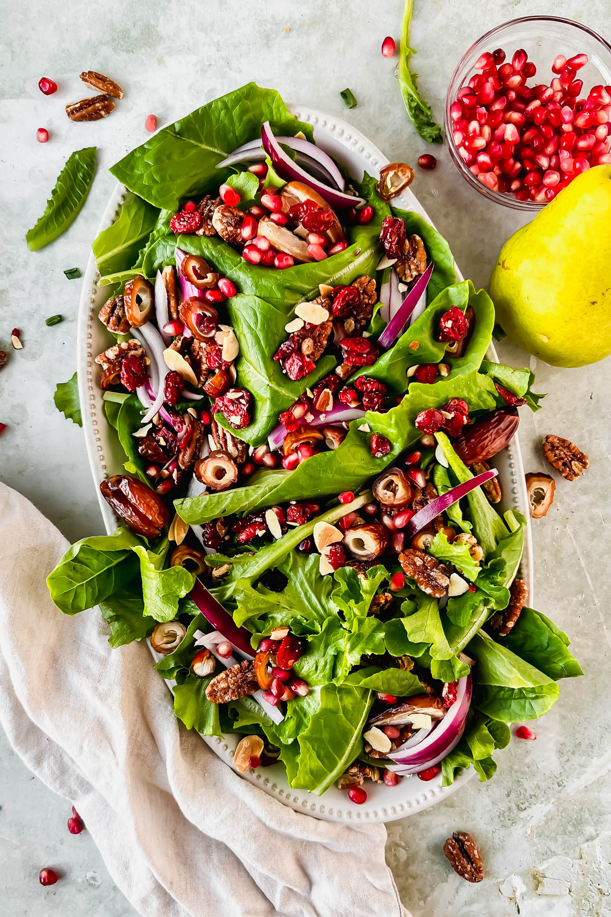 lettuce, sliced dates, pecans and red onion on white serving plate beside small bowl of pomegranate seeds and a pear.