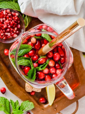 overhead shot of pomegranate champagne punch topped with fresh cranberries and mint leaves in large pitcher with ladle in pitcher.
