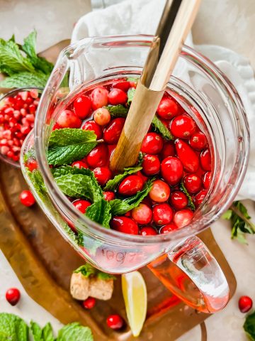overhead shot of pomegranate champagne punch topped with fresh cranberries and mint leaves in large pitcher with ladle in pitcher.
