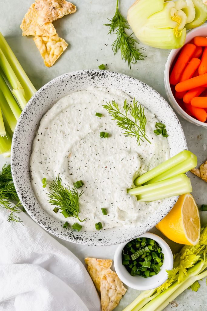 cottage cheese chip dip in speckled white bowl garnished with fresh dill and green onions with two celery sticks stuck in dish with chips and vegetables surrounding bowl.