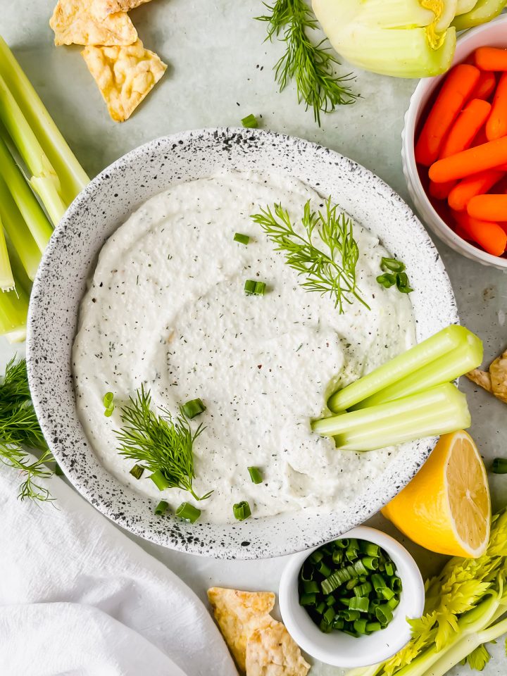 cottage cheese chip dip in speckled white bowl garnished with fresh dill and green onions with two celery sticks stuck in dish with chips and vegetables surrounding bowl.