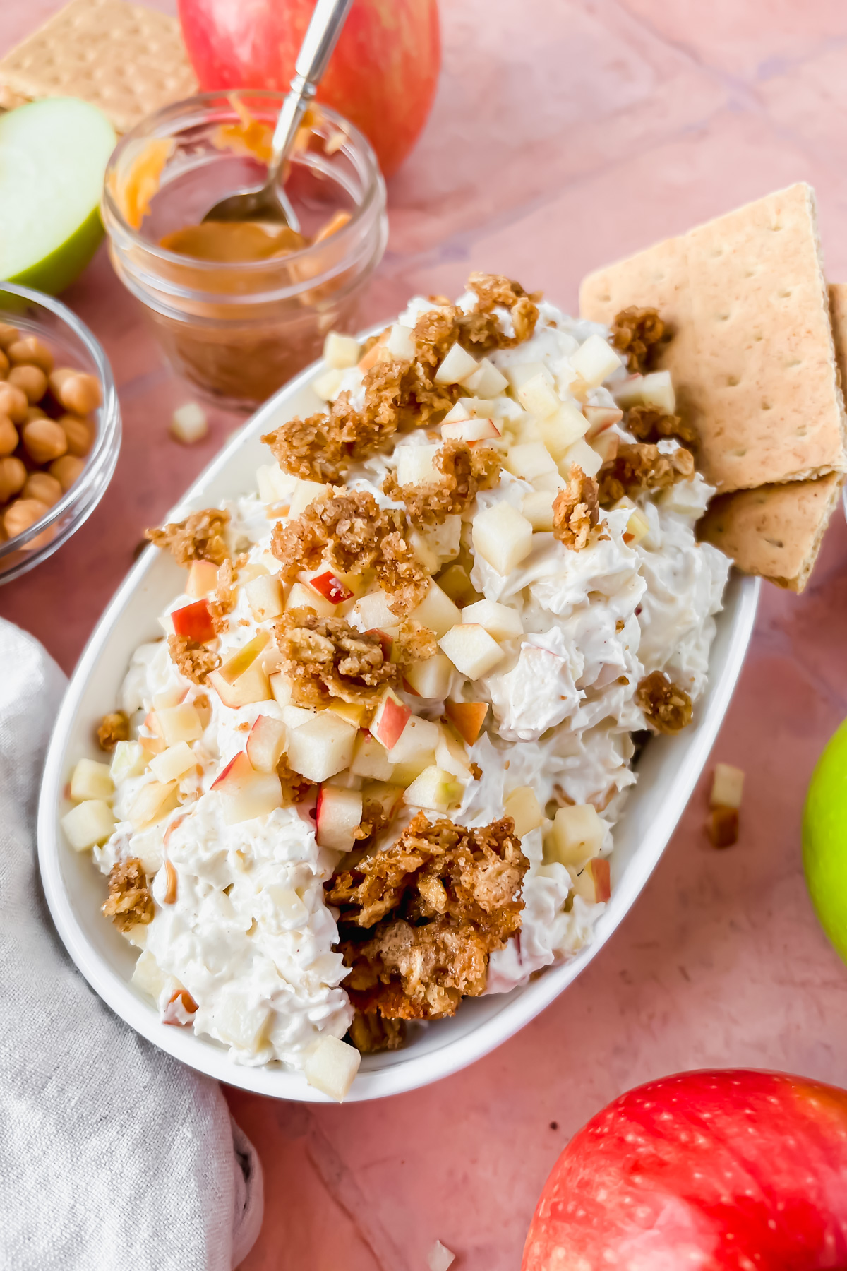 white oval serving dish filled with cream cheese caramel apple dip topped with granola, apple slices and two graham cracker squares surrounded by additional recipe ingredients.