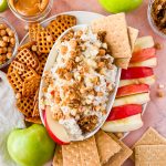 white oval serving dish filled with cream cheese caramel apple dip topped with granola, apple slices and two graham cracker squares surrounded by pretzels, graham crackers, and apples for dipping.