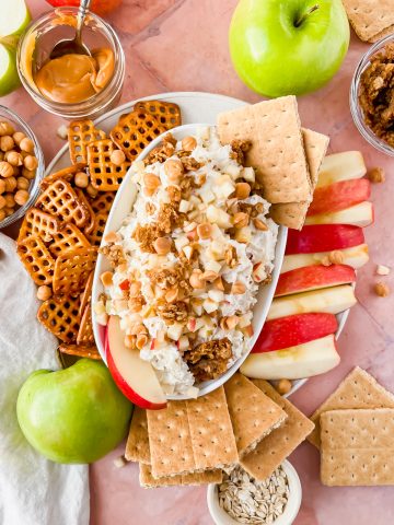 white oval serving dish filled with cream cheese caramel apple dip topped with granola, apple slices and two graham cracker squares surrounded by pretzels, graham crackers, and apples for dipping.