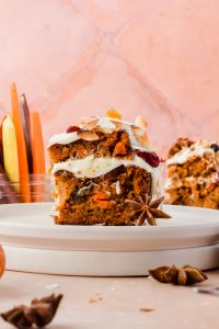 slice of two layer carrot cake on white plate with carrots and additional cake in background.