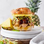 slow cooker pineapple pork on slider bun topped with extra barbecue sauce, broccoli slaw, and pineapple slice on white plate