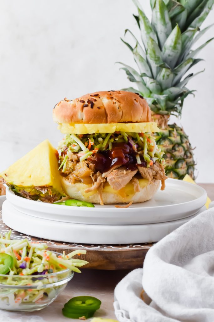 slow cooker pineapple pork on slider bun topped with extra barbecue sauce, broccoli slaw, and pineapple slice on white plate