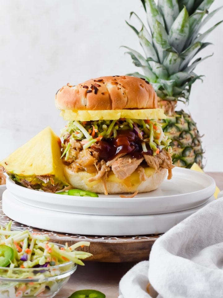 slow cooker pineapple pork on slider bun topped with extra barbecue sauce, broccoli slaw, and pineapple slice on white plate