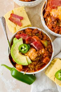 one serving of smokey bacon chili garnished with avocado and jalapeño slices beside slices of corn bread.