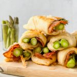 stacked asparagus in puff pastry with bacon on wood cutting board.