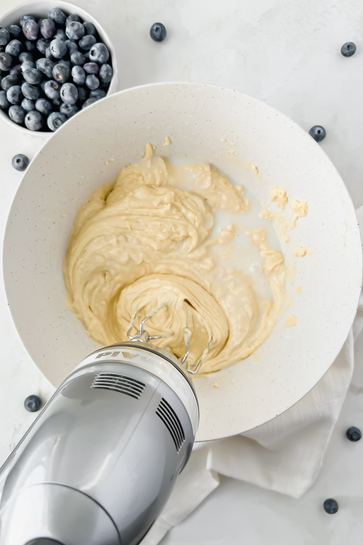 electric hand mixer mixing blueberry protein muffin batter in white mixing bowl.