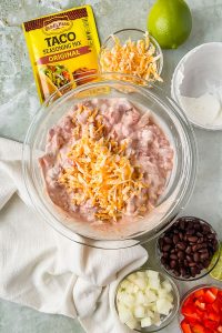 Chicken mixture for chicken taco casserole mixed in glass mixing bowl topped with shredded cheese and surrounded by additional recipe ingredients.