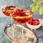 two cranberry fizz cocktail in stemmed cocktail glasses garnished with rosemary sprigs and fresh cranberries on silver tray.