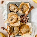 baked greek yogurt banana muffins baked in parchment liners arranged in a 6 muffin muffin tin with pecans, cinnamon sticks, and bananas surrounding.