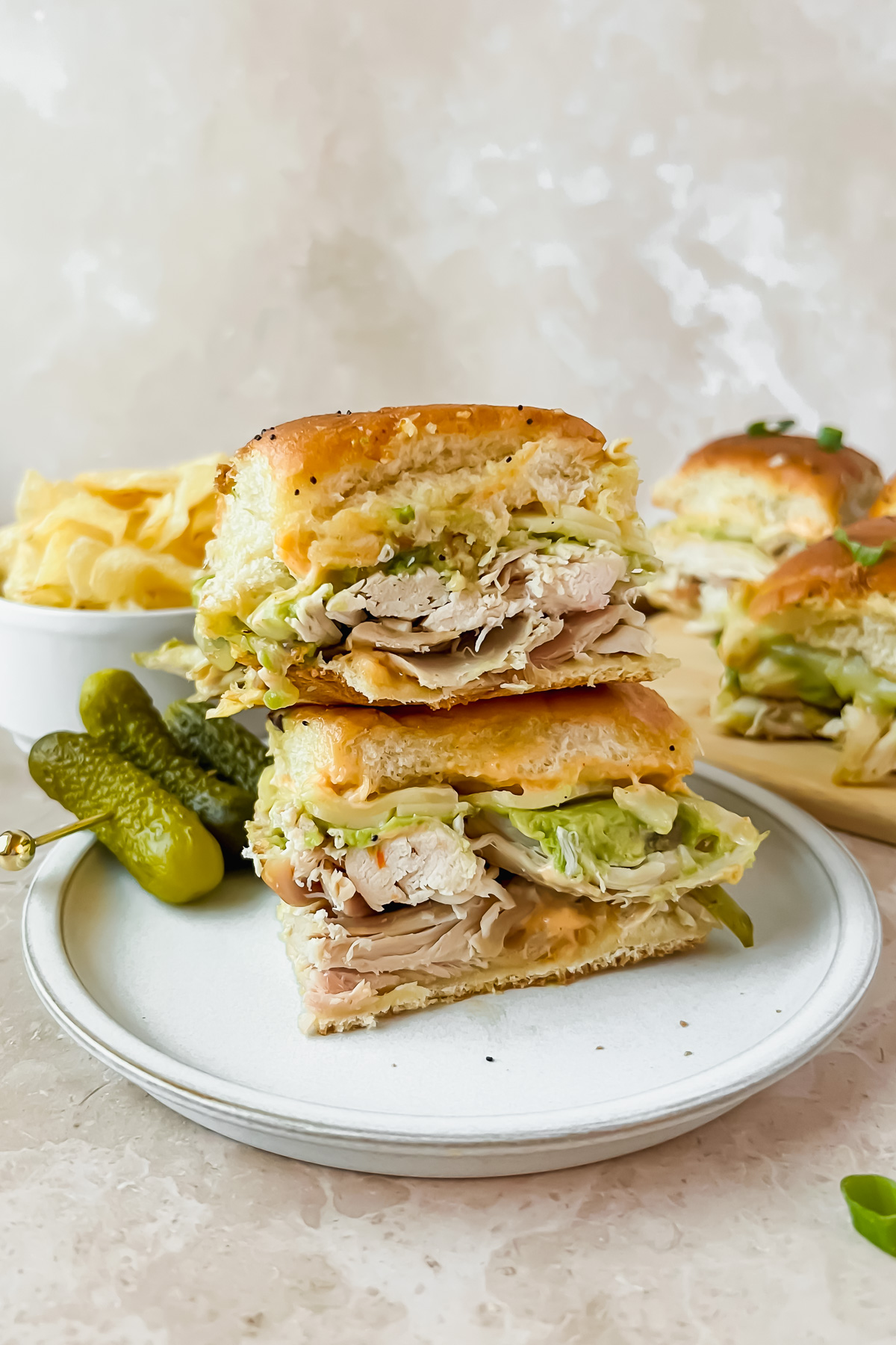 two stacked rotisserie chicken hawaiian sliders on white plate with bowl of chips and additional sliders in background.
