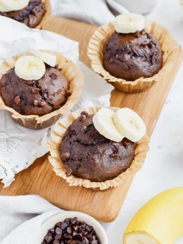 One Bowl Double Chocolate Banana Muffins topped with fresh banana slices on a wooden serving board.