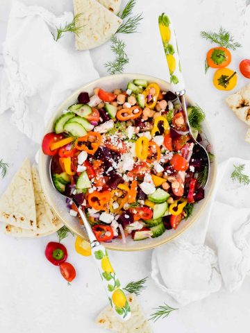 Mediterranean Chopped Salad in a white serving bowl with two serving spoons surrounded by mini peppers and pita bread.