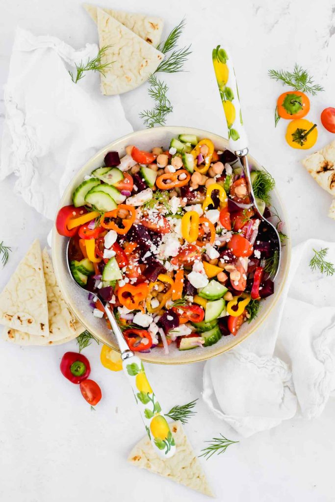 Mediterranean Chopped Salad in a white serving bowl with two serving spoons surrounded by mini peppers and pita bread.
