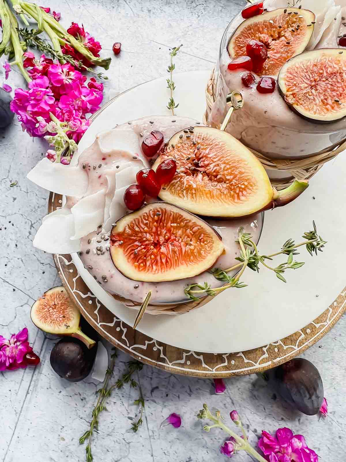 fig smoothie garnished with fresh fig halves, pomegranate seeds, and coconut flakes in glass with wicker sleeve on white tray with additional fig halves surrounding and vase of flowers in background.
