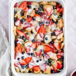 overhead shot of baked healthy french toast casserole garnished with fresh blueberries and strawberries with white glaze drizzled over top in white baking tray on white background.