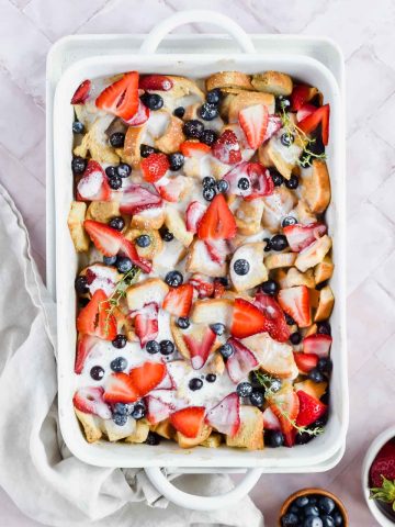 overhead shot of baked healthy french toast casserole garnished with fresh blueberries and strawberries with white glaze drizzled over top in white baking tray on white background.