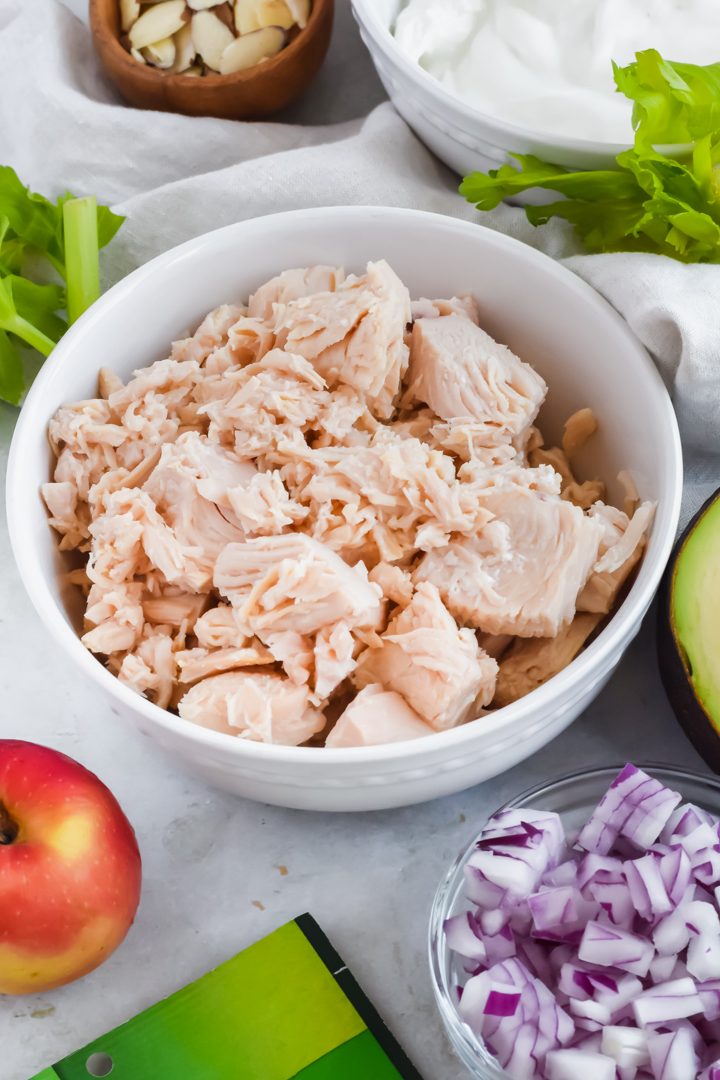 ingredients for pesto chicken salad including chicken, red onion, apple, avocado, almonds, and pesto in individual bowls.