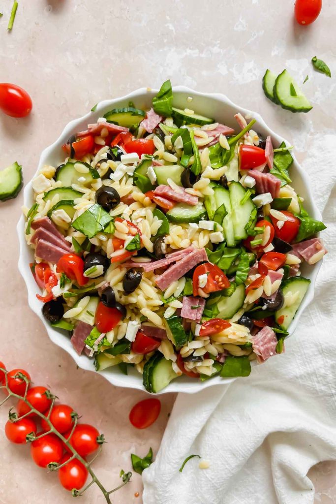one white bowl of italian orzo salad on stone background and assortment of vegetables around it.
