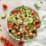 one white bowl of italian orzo salad on stone background and assortment of vegetables around it.