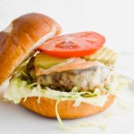 Oklahoma onion burger garnished with fresh tomato slice, pickles, and lettuce on brioche bun on white plate.