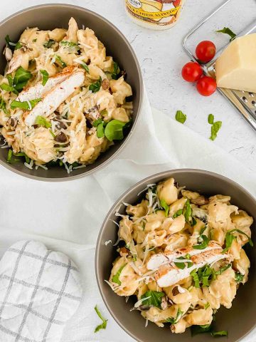 two bowls of creamy alfredo pasta garnished with sliced chicken, parmesan, and greens.