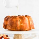 hand pouring frosting on peach bundt cake with fresh peaches surrounding it.