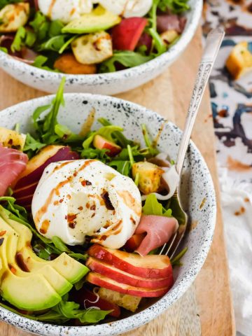 white ceramic salad bowl filled with arugula, peaches, avocado slices, and burrata cheese with balsamic dressing.