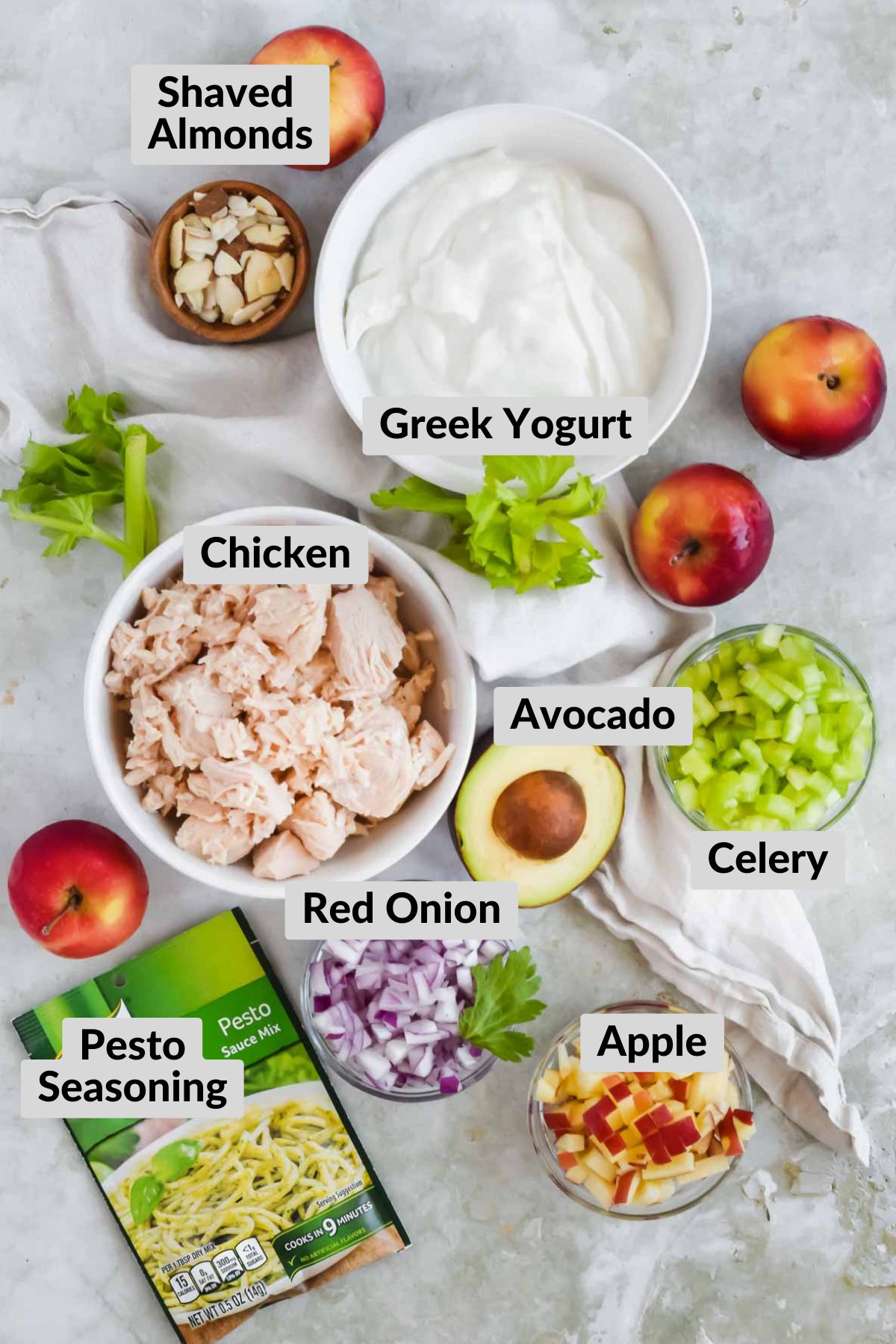 ingredients for pesto chicken salad including chicken, red onion, apple, avocado, almonds, dressing, celery, and pesto seasoning.