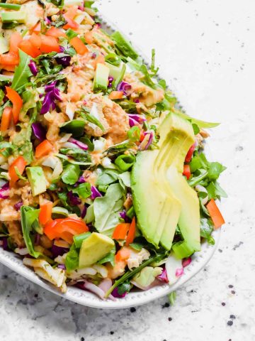 close up of spicy thai chicken salad garnished with peanut dressing and fresh avocado slices on white serving dish with speckled white background.