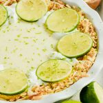 half of a tequila key lime pie garnished with limes and toasted coconut on white background.