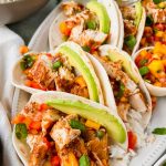 blackened mahi tacos with mango salsa and avocado on white serving plate surrounded by taco toppings.