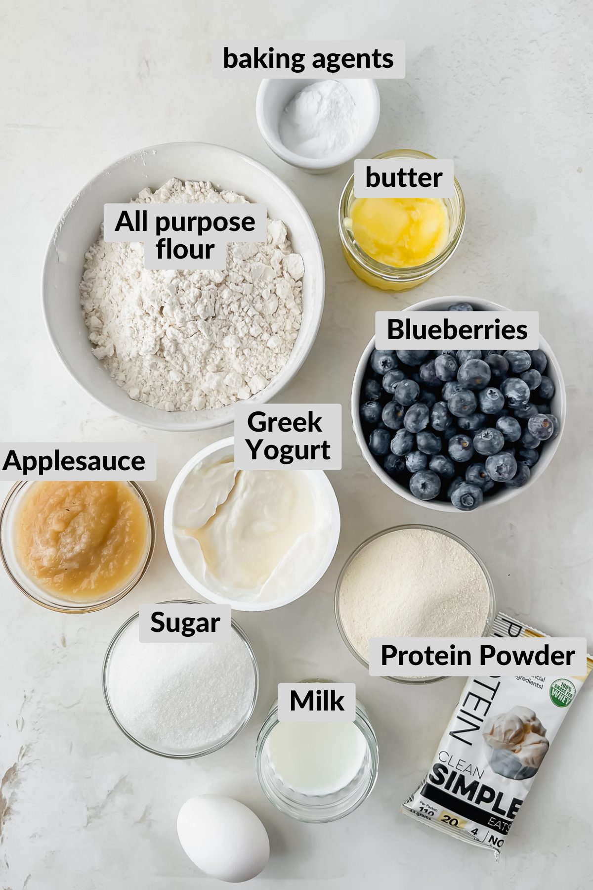 ingredients for blueberry protein muffins in individual bowls arranged on white background.