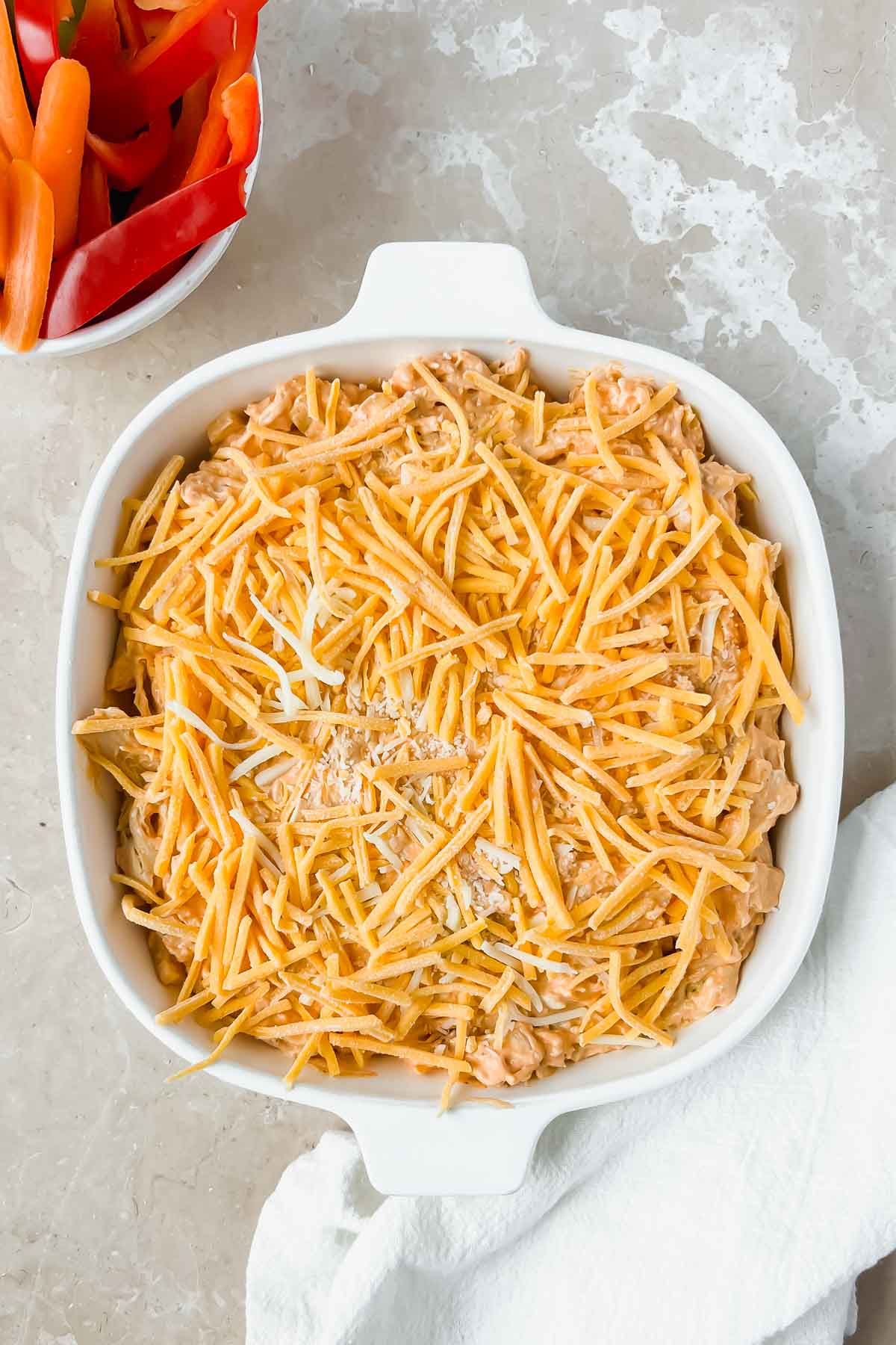 metal square pan filled with uncooked buffalo chicken dip topped with shredded cheese on white background.