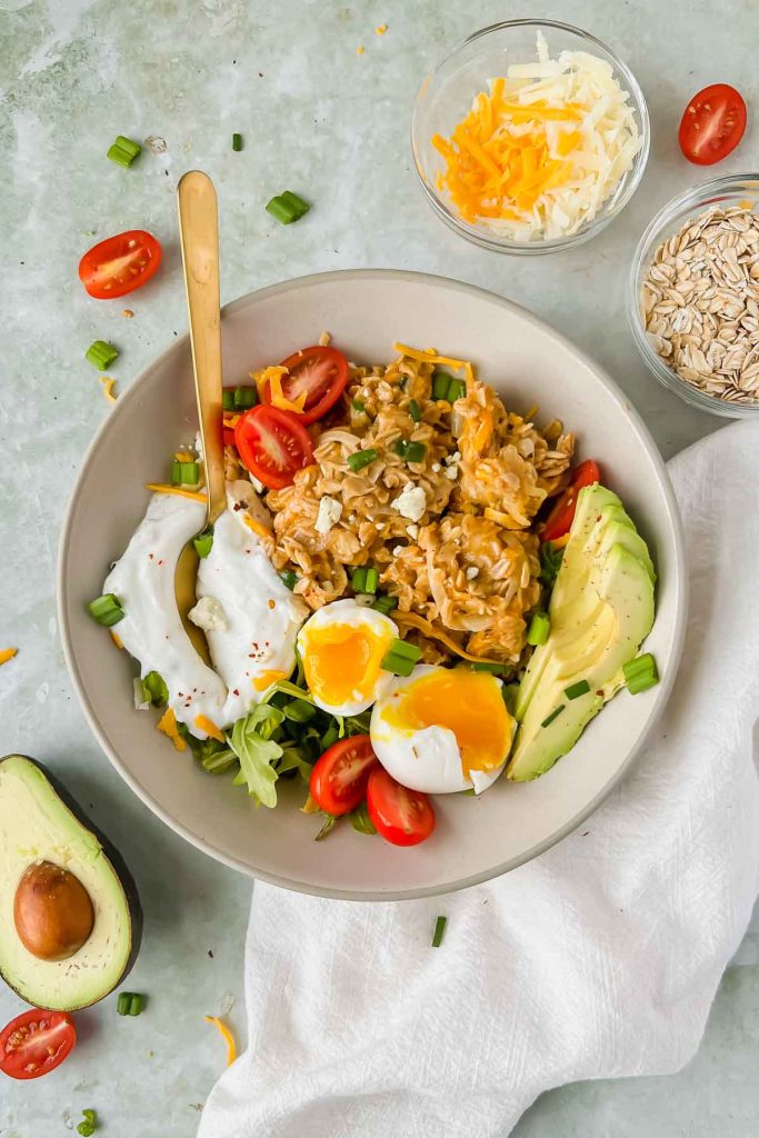 cheesy oatmeal topped with avocado, egg, yogurt, and herbs with a gold spoon in a ceramic bowl on a gray slate background.