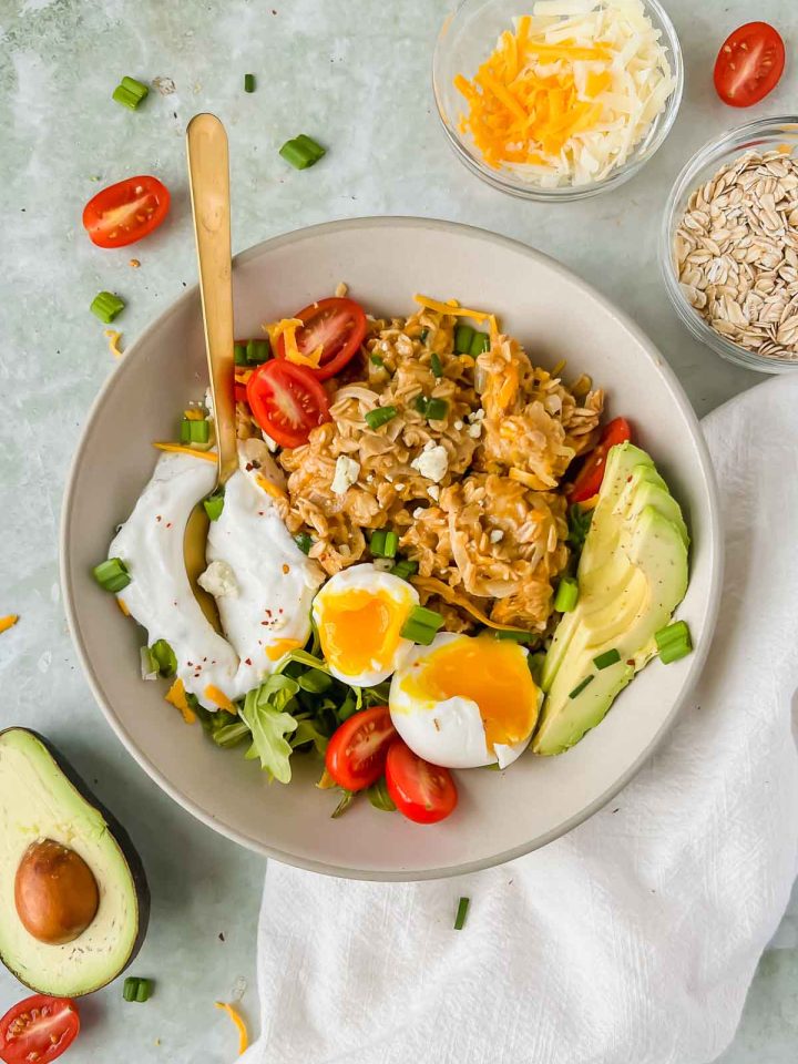 cheesy oatmeal topped with avocado, egg, yogurt, and herbs with a gold spoon in a ceramic bowl on a gray slate background.