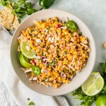 mexican street corn dip served in a grey ceramic bowl garnished with lime wedges, cilantro leaves, on a green backdrop.