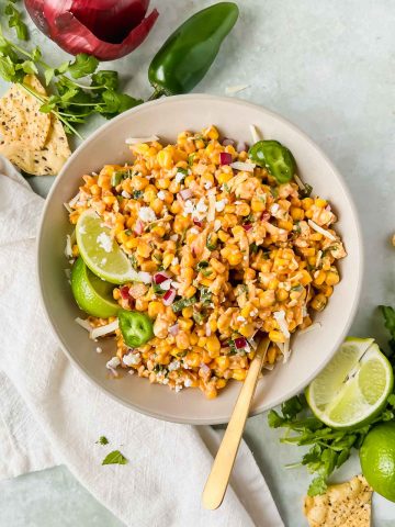 gold spoon inserted into mexican street corn dip served in a grey ceramic bowl garnished with lime wedges, cilantro leaves, on a green backdrop.