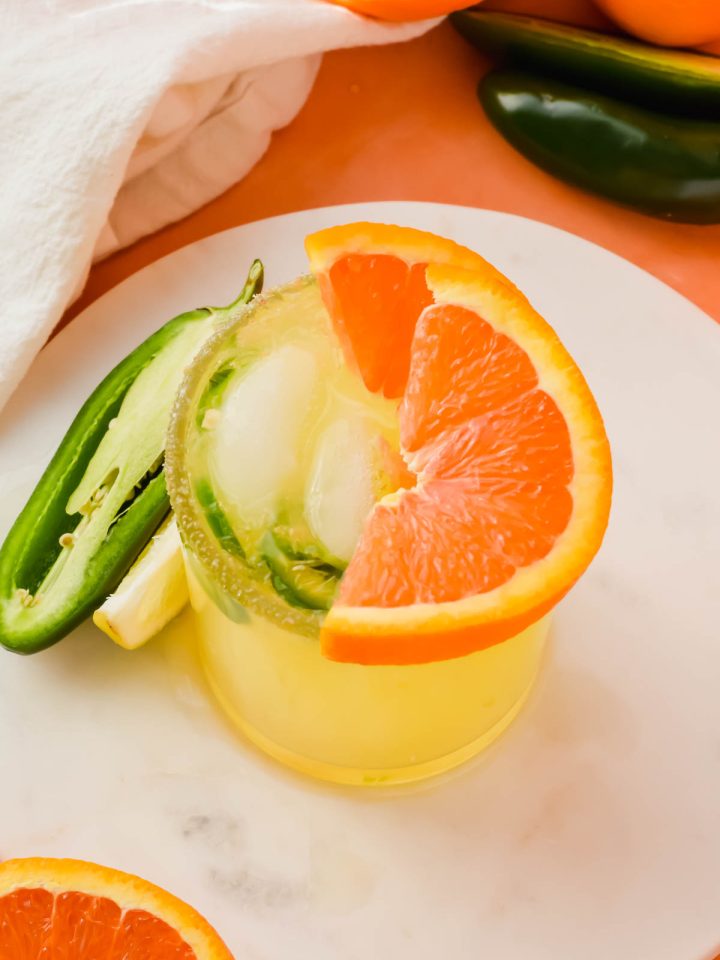 spicy orange margarita garnished with jalapeno slices and oranges on a white marble slab.