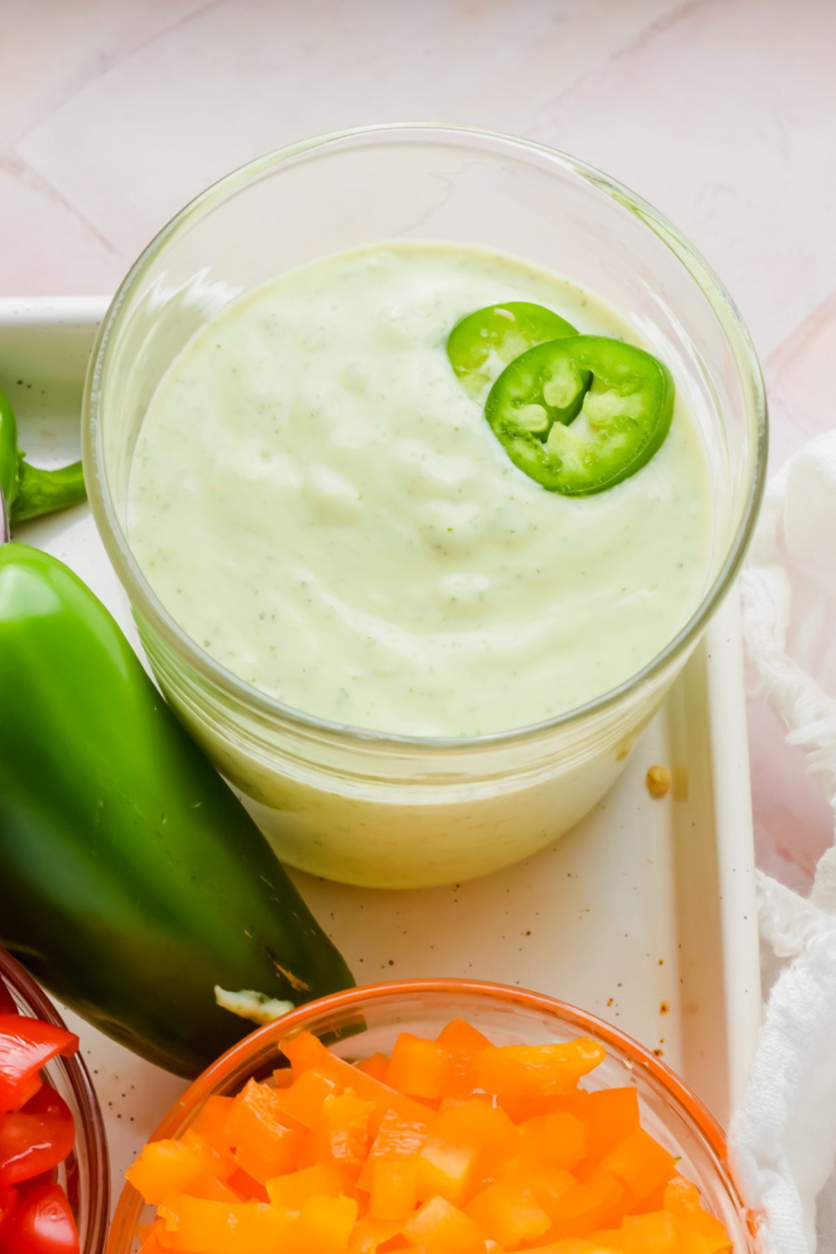 creamy jalapeno sauce in glass beaker beside fresh jalapeno and small bowls of chopped peppers, tomatoes, and onion.