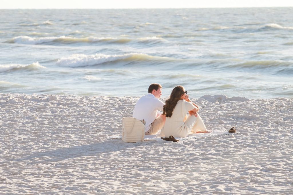 man and woman sitting on a cream blanket in the sand staring at the ocean. 
