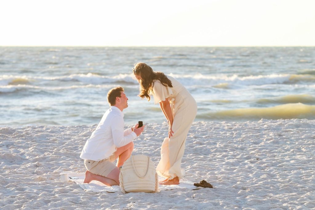 man bending down on one knee proposing to girl standing in cream jumpsuit on a beach at sunset.