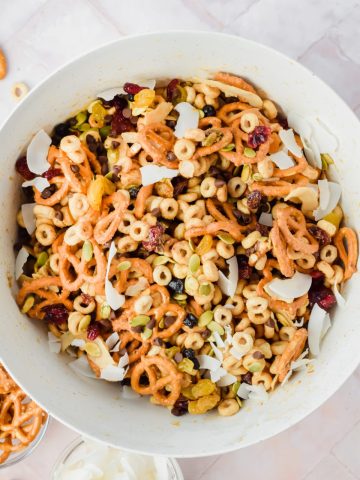cheerio trail mix in large white bowl beside two smaller bowls containing pretzels and coconut flakes.