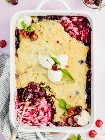 white baking dish filled cherry blueberry cobbler with a spoon inserted into corner of dish.
