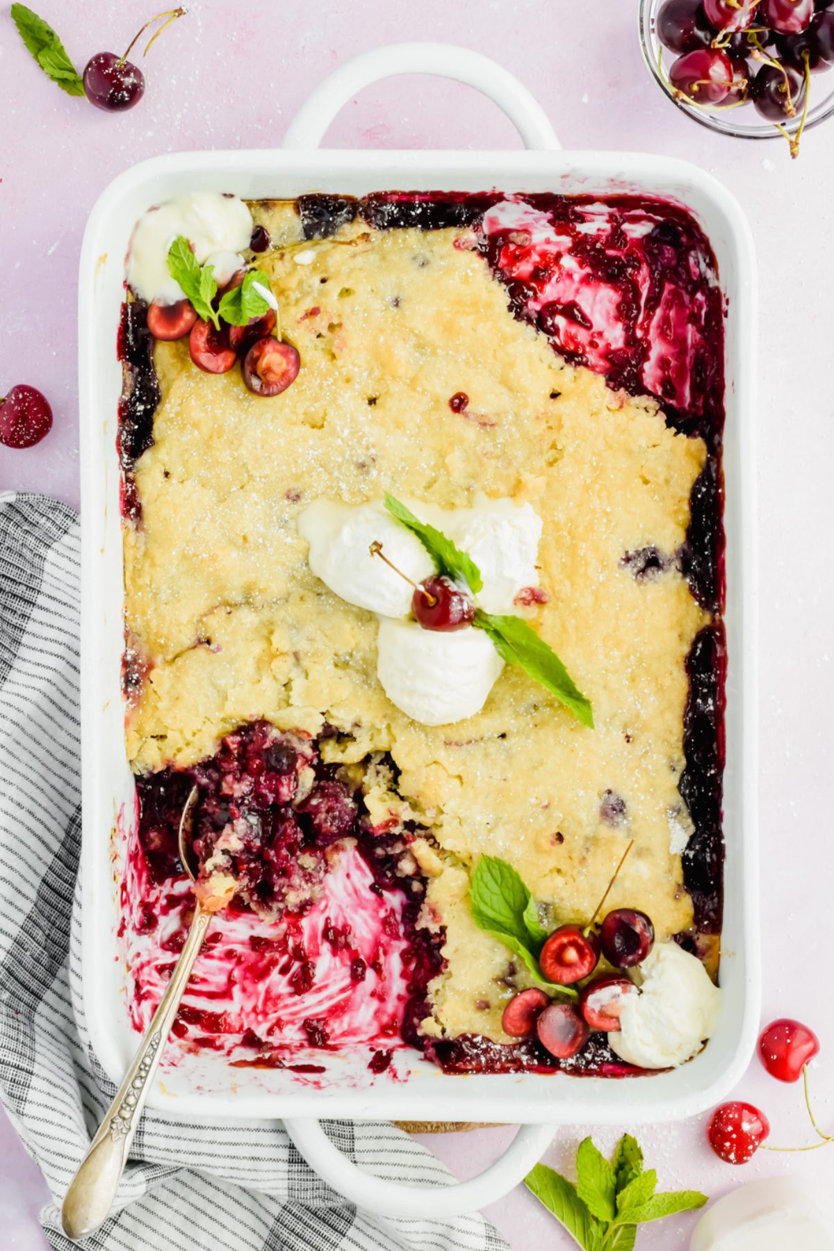 white baking dish filled cherry blueberry cobbler with a spoon inserted into corner of dish.